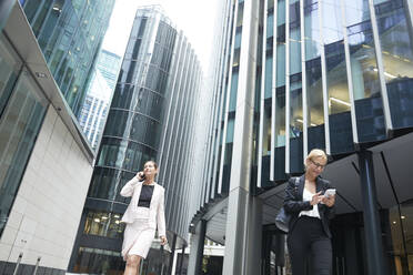 Female professionals walking with mobile phones against office building at downtown district in city - PMF01301