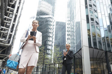 Mature businesswomen using smart phones against modern office building at downtown district - PMF01304