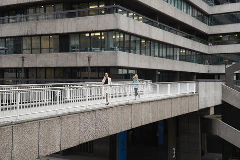 Frau, die an einer weiblichen Fachkraft vorbeiläuft, die auf einer Fußgängerbrücke im Stadtzentrum mit einem Mobiltelefon spricht - PMF01308