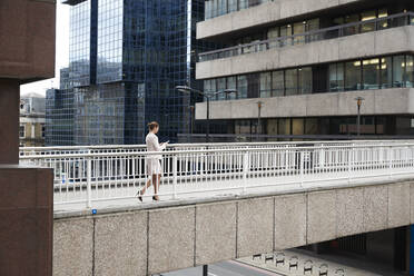 Geschäftsfrau, die ein Smartphone benutzt, während sie auf einer Fußgängerbrücke im Stadtzentrum spazieren geht - PMF01309