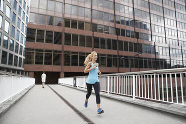 Mature blond woman jogging against businesswoman on footbridge in financial district - PMF01314