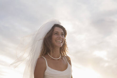 Cheerful young bride looking away while standing at beach against sky during sunset - AJOF00185