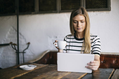 Blonde Frau, die ein digitales Tablet benutzt und Kaffee trinkt, während sie in einem Cafe sitzt - ABZF03405