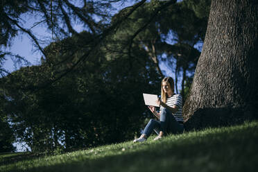 Woman using digital tablet while sitting on grass in public park - ABZF03414