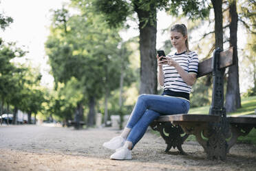 Frau schreibt Textnachrichten auf ihrem Smartphone, während sie in einem öffentlichen Park sitzt - ABZF03417