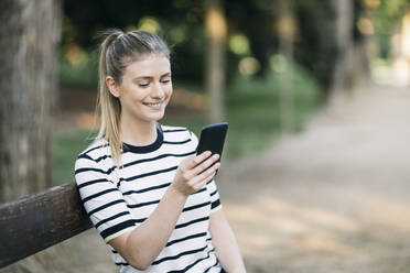 Frau lächelt, während sie in einem öffentlichen Park eine Textnachricht auf ihrem Smartphone verschickt - ABZF03418