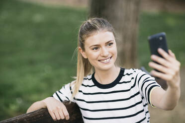 Beautiful woman taking selfie on smart phone while sitting in public park - ABZF03419