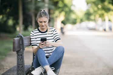 Mittlere erwachsene Frau, die in einem öffentlichen Park sitzt und ihr Smartphone benutzt - ABZF03420