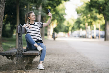 Frau, die ein Selfie mit ihrem Smartphone macht, während sie in einem öffentlichen Park sitzt - ABZF03421