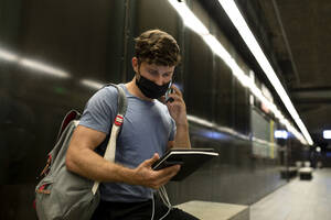 Young man wearing protective face mask talking on phone while holding digital tablet at subway during pandemic - VABF03587