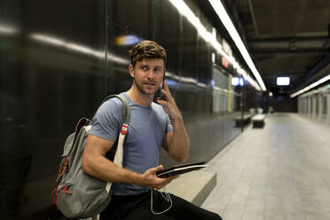 Gut aussehender junger Mann, der telefoniert und ein digitales Tablet hält, während er auf eine beleuchtete U-Bahn blickt - VABF03588