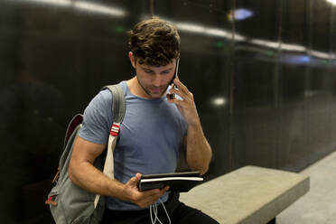 Junger männlicher Pendler, der in einer beleuchteten U-Bahn an der Wand sitzend telefoniert und ein digitales Tablet hält - VABF03589