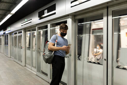 Junger männlicher Pendler, der sein Smartphone benutzt, während er in einem fahrenden Zug in einer U-Bahn-Station steht, während COVID-19 - VABF03591