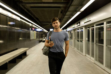 Gut aussehender junger Mann, der sein Smartphone in der Hand hält, während er mit der Hand in der Tasche auf einem beleuchteten U-Bahn-Bahnsteig geht - VABF03593
