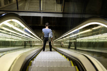 Young man with backpack standing on illuminated escalator at subway - VABF03595