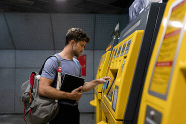 Gutaussehender junger männlicher Pendler, der ein Buch hält, während er einen Fahrkartenautomaten am Bahnhof benutzt - VABF03600