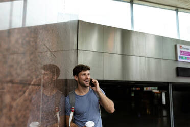 Lächelnder Mann, der durch sein Smartphone spricht, während er wegschaut und sich an eine braune Kachelwand in einer U-Bahn-Station lehnt - VABF03604