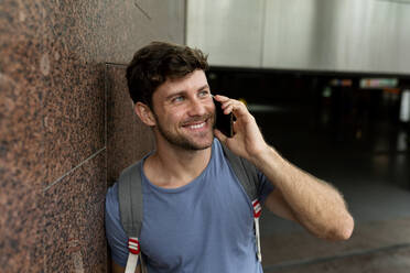 Glücklicher Mann, der mit seinem Handy spricht, während er wegschaut und sich an eine braune Kachelwand in einer U-Bahn-Station lehnt - VABF03605