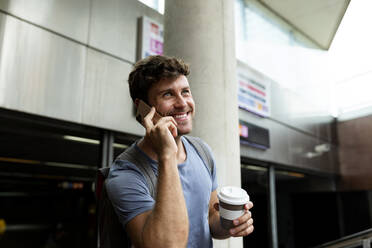 Lächelnder junger Pendler, der mit seinem Smartphone spricht, während er wegschaut und einen wiederverwendbaren Kaffeebecher in der U-Bahn-Station hält - VABF03606