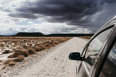 Spain, Navarre, Storm clouds over car driving along empty dirt road in Bardenas Reales - EBBF00835