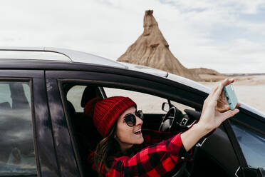 Spain, Navarre, Female tourist leaning out of car window taking smart phone photos of Bardenas Reales - EBBF00852