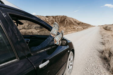 Spain, Navarre, Young woman sticking legs out of car window over over dirt road in Bardenas Reales - EBBF00854