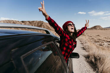 Spain, Navarre, Female tourist leaning out of car window making peace sign gestures over dirt road in Bardenas Reales - EBBF00856