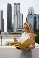 Blonde Frau analysiert ein Dokument in einer Akte, während sie vor Wolkenkratzern im Stadtzentrum steht - MAUF03541