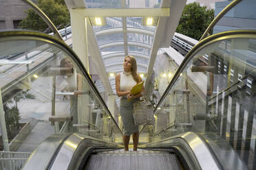 Nachdenkliche blonde Frau mit Blick nach oben, während sie auf einer Rolltreppe in der Stadt steht - MAUF03547