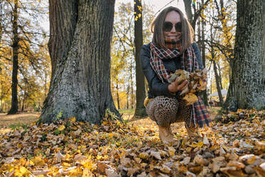 Smiling woman crouching while holding dry leaves against trees at park during autumn - KNTF05682