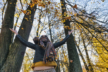 Frau wirft trockene Herbstblätter, während sie mit ausgestreckten Armen gegen Bäume im Park steht - KNTF05684