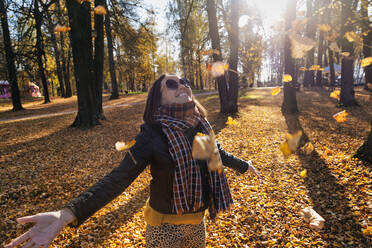 Smiling beautiful woman with arms outstretched enjoying while standing under falling dry leaves at park during autumn - KNTF05686