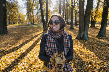 Lächelnde schöne Frau, die trockene Blätter hält, während sie im Herbst im Park steht - KNTF05687