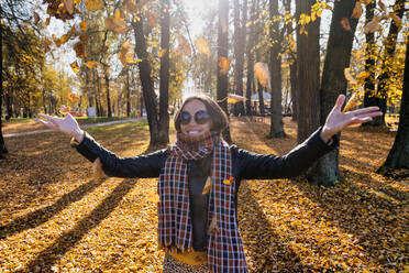 Lächelnde schöne Frau mit ausgestreckten Armen, die trockene Blätter wirft, während sie in einem Park im Herbst steht - KNTF05688