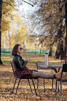 Mid adult woman drinking coffee while sitting in public park - KNTF05694