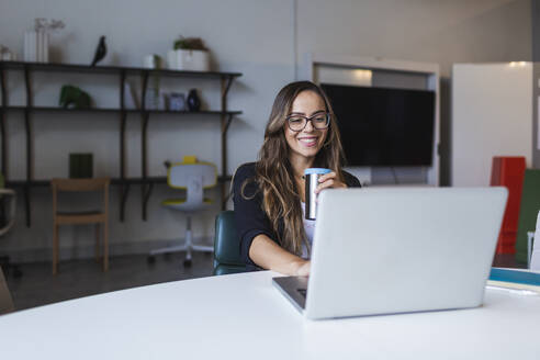 Lächelnde Geschäftsfrau, die Kaffee trinkt und einen Laptop am Schreibtisch im Büro benutzt - DCRF00927