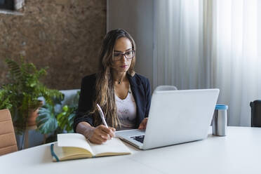 Creative businesswoman using laptop while writing in diary at office - DCRF00928