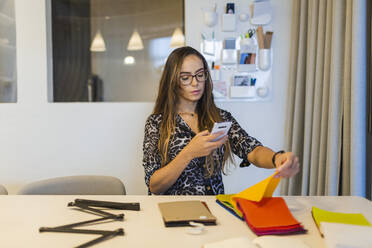Kreative Geschäftsfrau, die mit ihrem Smartphone ein Stoffmuster fotografiert, während sie am Schreibtisch im Büro sitzt - DCRF00933