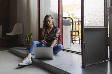 Unternehmerin, die ein Smartphone und einen Laptop benutzt, während sie in einem kreativen Büro an der Tür sitzt - DCRF00936