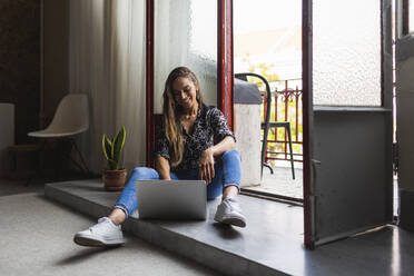 Smiling creative businesswoman working on laptop while sitting at doorway in office - DCRF00938