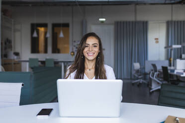 Lächelnde Geschäftsfrau mit Laptop am Schreibtisch im Büro - DCRF00946