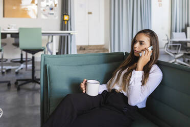 Geschäftsfrau, die mit dem Handy telefoniert, während sie auf dem Sofa im Büro Kaffee trinkt - DCRF00949