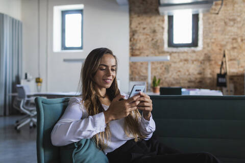 Smiling businesswoman text messaging through mobile phone while sitting on sofa in office stock photo