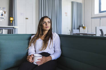 Nachdenkliche Geschäftsfrau mit Kaffeetasse auf dem Sofa im Büro - DCRF00951
