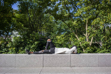 Man with cap on face lying on retaining wall at park - VPIF03071