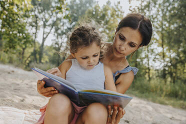 Mutter und Tochter lesen ein Buch, während sie am Strand sitzen - MFF06234