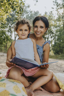 Glückliche Mutter und Tochter lesen ein Buch, während sie am Strand sitzen - MFF06236