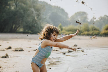 Cute girl throwing sand and mud while standing at beach - MFF06258