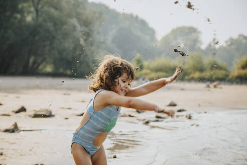 Nettes Mädchen wirft Sand und Schlamm, während sie am Strand steht - MFF06258