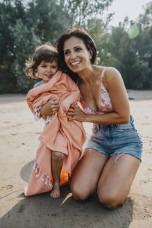 Lächelnde Mutter schaut weg, während sie ihrer Tochter am Strand ein Handtuch umlegt - MFF06264